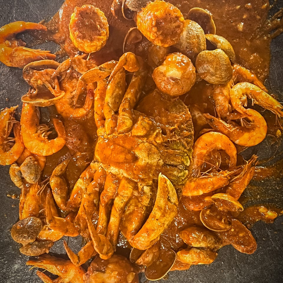 Crab, shrimp, clams, and corn on the cob covered in a spicy reddish-brown sauce on a dark surface.